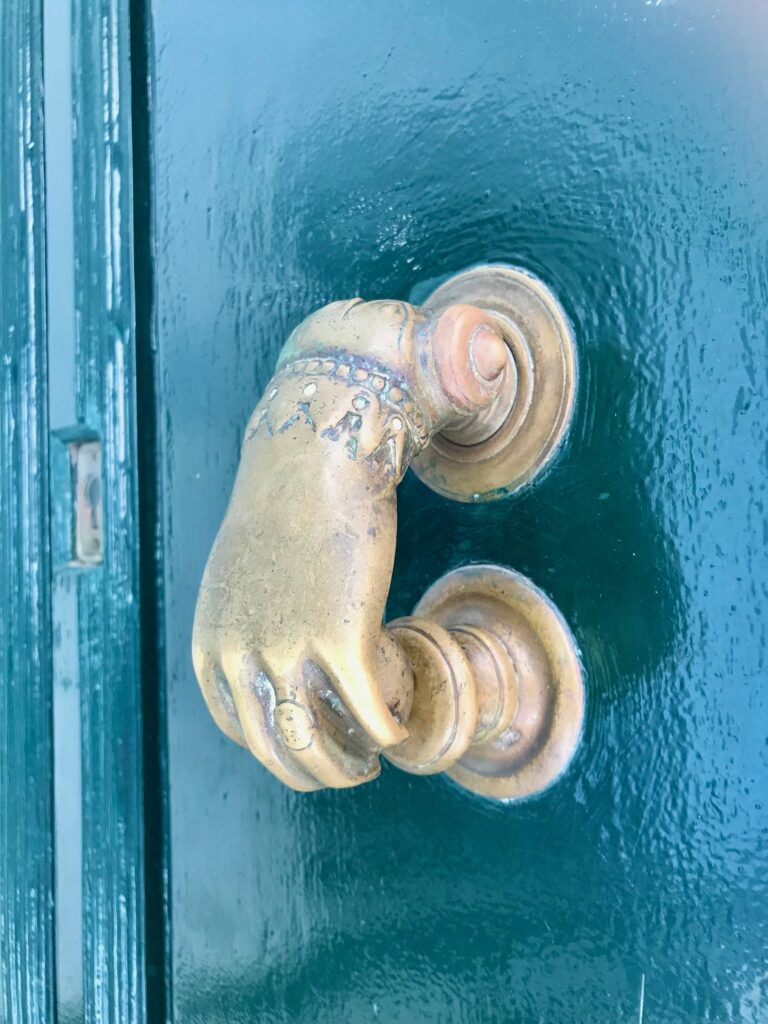 En bild på en antik dörrkläpp i mässing föreställande en kvinnohand på en blåmålad dörr i trä.